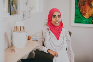 Photo of Fariha Tayyab wearing a red headscarf and white coat, leaning against a black chair with a green and brown painting behind her. 